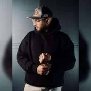 Confident man in a black hoodie with 'URBAN SQUAD JUST FEEL IT' embossed text, wearing a beige and gray cap and a stylish watch, adjusting his sleeve.